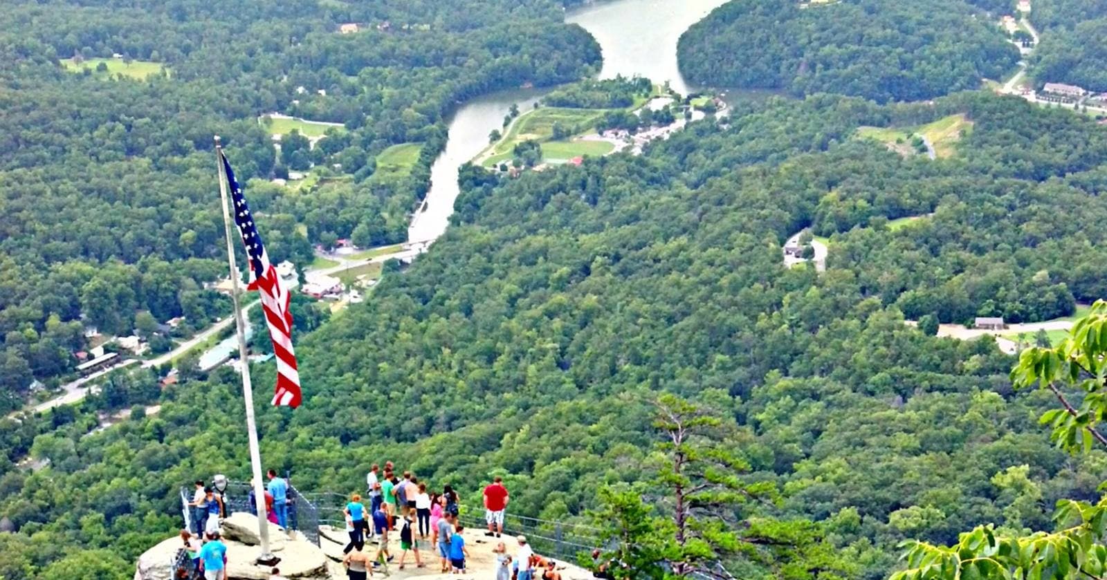 Visitors on the Chimney Rock summit observation deck beside the American flag, with Lake Lure and the Rocky Broad River valley below