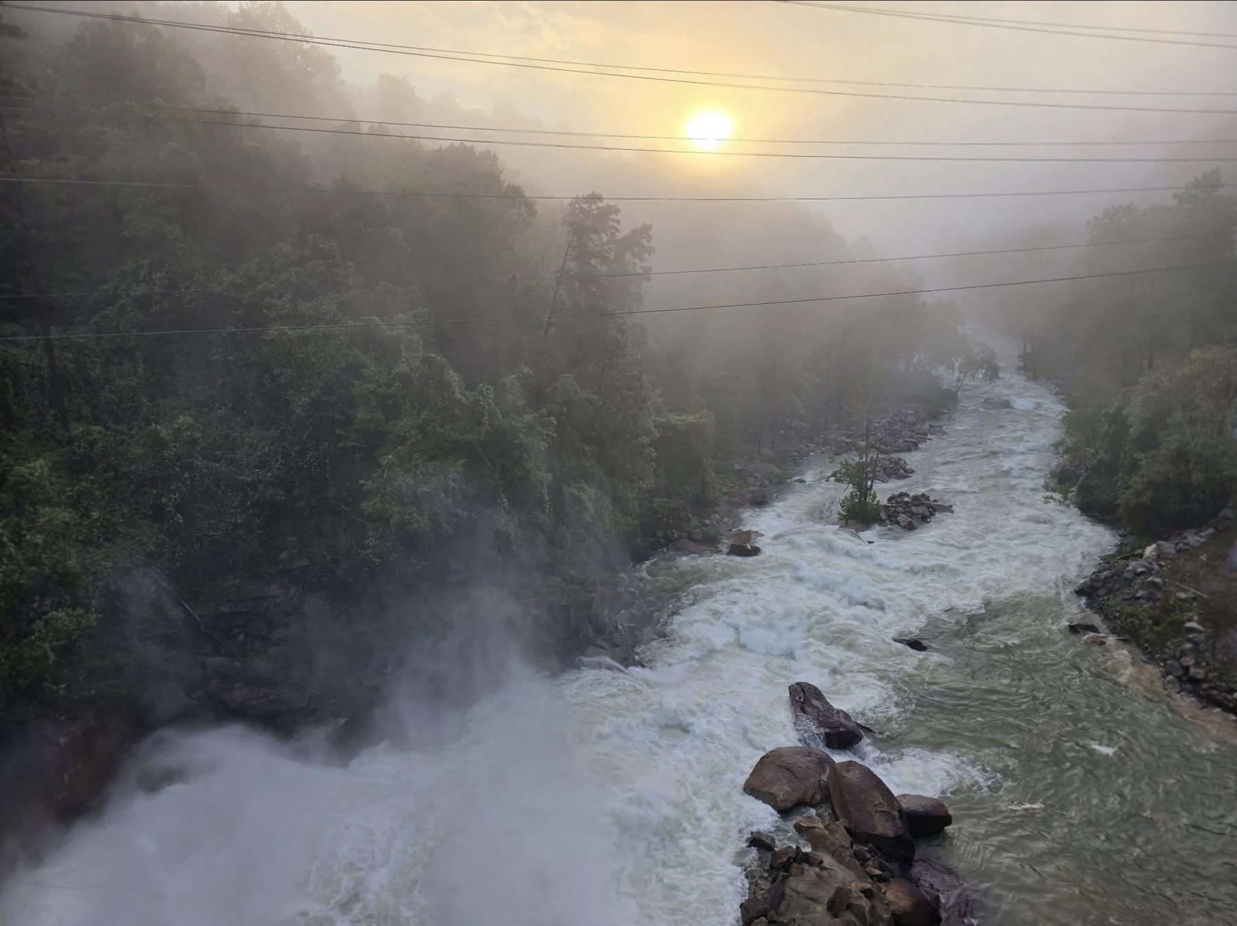 Fast, churning whitewater and mist in the Broad River channel just below Lake Lure Dam at sunrise on Sunday, Oct. 11, 2026, with fog on the wooded hillsides and power lines crossing overhead.